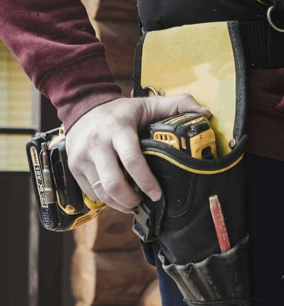 Electrician's hand reaching for a cordless drill in a tool belt, showcasing professional equipment for domestic electrical work.