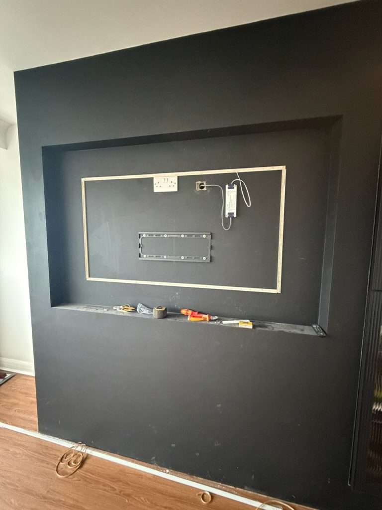 Electrical installation setup on a black wall featuring a mounted frame for a TV or display, with visible wiring, power outlet, and tools on the floor, illustrating DB Electrics' services for new build and extension electrical installations in Maidenhead.
