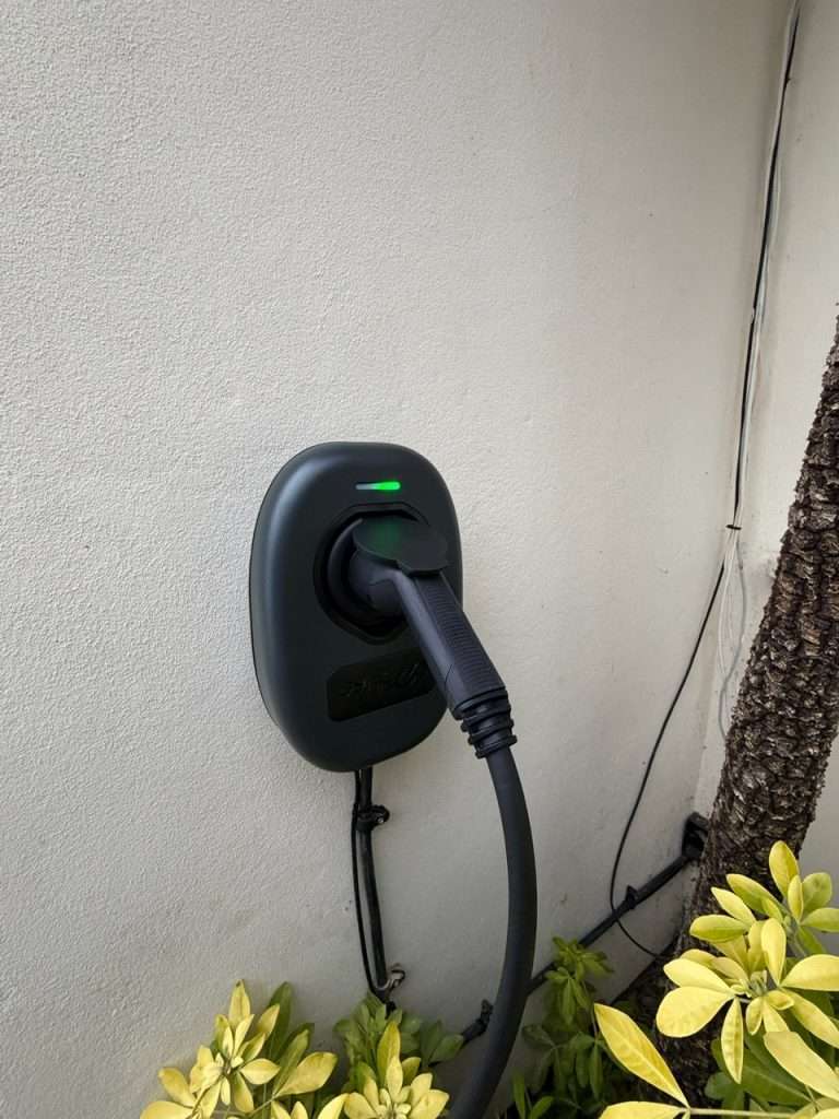 Wall-mounted 7kW EV charger with charging cable connected, installed on a wall near green foliage, indicating home electric vehicle charging solution in Maidenhead.