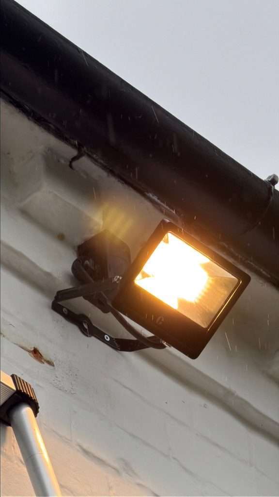 Outdoor floodlight installation on a white wall, illuminating the area, with rain droplets visible, showcasing weatherproof lighting solutions by DB Electrics for garden and outdoor electrical installations in Maidenhead.